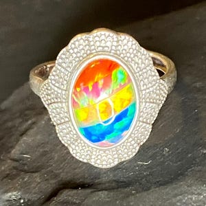 Op de afbeelding: Zilveren ring met een ovale, iriserende edelsteen met levendige kleuren rood, oranje, geel, groen en blauw. De edelsteen is gezet in een gedetailleerde, geschulpte bezel.