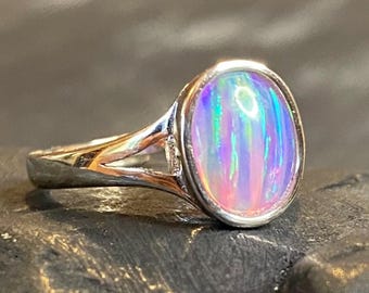 May include: A silver ring with an oval opal gemstone. The opal displays a vibrant play of color, including pink, blue, and green hues. The ring's band has a split design, adding a unique detail to the jewelry.