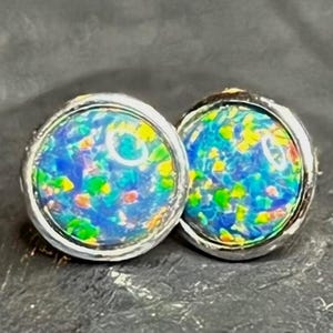 May include: Two round stud earrings with a silver-coloured setting. Each earring features a vibrant, multicoloured opal-like stone with shades of blue, green, yellow, and red. The stones have a glossy finish, and the earrings are set against a dark background.