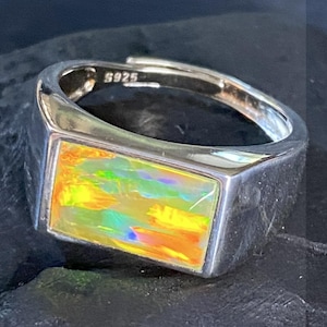 Puede incluir: Un anillo de plata con una piedra de ópalo rectangular. El ópalo muestra una vibrante gama de colores que incluyen amarillo, naranja, verde y azul. El anillo está estampado con "S925".