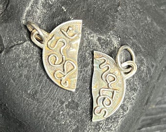 May include: Two silver-toned pendants, each shaped like a half-circle, with a loop at the top for a chain. One pendant reads "Love" and the other reads "Mates" in a cursive font. The pendants are on a dark, textured surface.