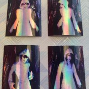Op de afbeelding: Vier iriserende holografische stickers met een figuur in een witte gewatteerde kap met een donkere achtergrond.