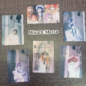 May include: Six glossy photo cards featuring a Japanese visual kei band called Malice Mizer. The cards are white with black text and feature images of the band members in various poses.