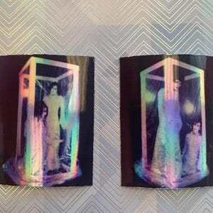 Op de afbeelding: Twee iriserende holografische afbeeldingen van mensen die in een kubusvormig frame staan. De afbeeldingen zijn op een zwarte achtergrond.