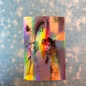 May include: A holographic sticker featuring a woman with dark hair and a white bow, standing in a doorway with a rainbow effect. The sticker says "BUTKI" in bold, black letters.