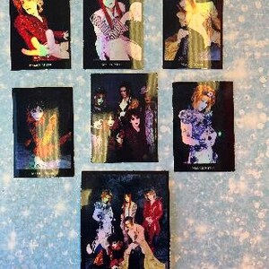 May include: A set of six black and white holographic trading cards featuring a Japanese rock band. The cards feature different band members in various poses. The band's name is printed on the bottom of each card.