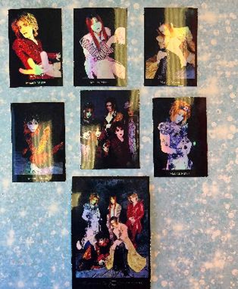 May include: A set of six black and white holographic trading cards featuring a Japanese rock band. The cards feature different band members in various poses. The band's name is printed on the bottom of each card.