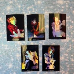 May include: Set of five holographic trading cards featuring a person with long, dark hair wearing a variety of colorful outfits. The cards have a black background and feature the text "Mana Star" on the bottom.