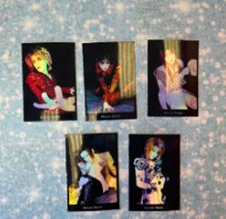 May include: Set of five holographic trading cards featuring a person with long, dark hair wearing a variety of colorful outfits. The cards have a black background and feature the text "Mana Star" on the bottom.