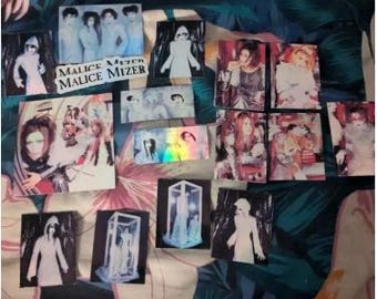 Puede incluir: Una colección de 14 pegatinas con imágenes de una banda de rock japonesa, Malice Mizer. Las pegatinas están impresas en papel blanco y tienen un acabado brillante. Algunas de las pegatinas muestran a los miembros de la banda con sus característicos abrigos blancos con capucha, mientras que otras muestran imágenes de tigres y otros animales. Las pegatinas son de diferentes tamaños y formas.