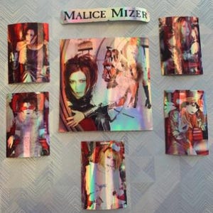 Op de afbeelding: Een set van zeven holografische stickers met de band Malice Mizer. De stickers zijn rechthoekig en bevatten verschillende afbeeldingen van de bandleden. De stickers zijn gerangschikt in een rasterpatroon op een witte achtergrond.