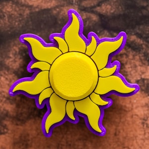 May include: A yellow sun-shaped charm with a purple outline. The sun has a textured center and curved rays. This decorative charm is likely for a bracelet or necklace.