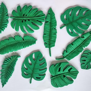 May include: A collection of green, artificial tropical leaves, including monstera and palm leaves, arranged on a white surface. The leaves vary in size and shape, with detailed veining and textures.