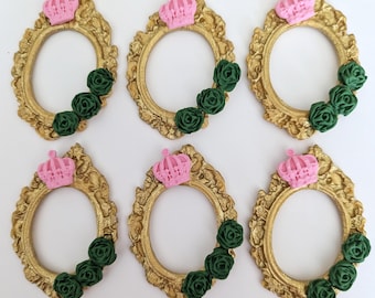 May include: Six gold-colored oval frames with ornate detailing. Each frame is adorned with a pink crown at the top and a cluster of three dark green roses at the bottom. The frames are arranged on a white surface.