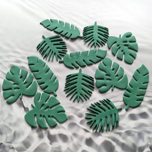 May include: A collection of green, stylized leaf cutouts in various shapes and sizes. The leaves are arranged on a white and gray textured background, creating a tropical or botanical theme. The leaves have detailed veining.