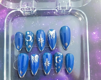 May include: A set of ten blue press-on nails with silver celestial designs, including moons, stars, and sunbursts. The nails are oval-shaped and have a silver metallic trim. They are displayed in a clear plastic case.