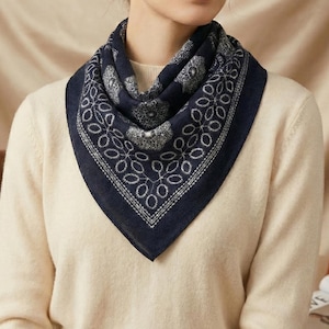 May include: A navy blue scarf with a white floral pattern and a white border is draped around the neck. The scarf is worn over a cream-colored sweater. The scarf's design is symmetrical and geometric, creating a classic look.