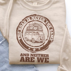 Puede incluir: Sudadera beige de cuello redondo con un gráfico circular marrón. El gráfico presenta una furgoneta vintage con equipaje en la parte superior, rodeada por el texto "THE ROAD IS NEVER THE SAME" y "AND NEITHER ARE WE".