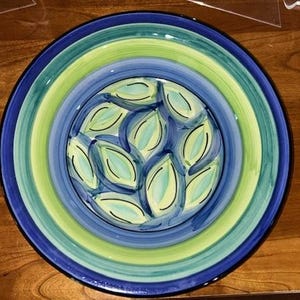 May include: A round ceramic plate with a hand-painted design. The plate features concentric rings of blue, green, and yellow. The center has a pattern of blue leaves on a white background. The plate is on a wooden surface.