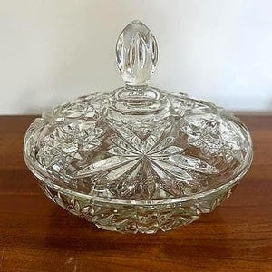 May include: A clear, cut-glass candy dish with a lid. The dish has a starburst pattern on the base and lid, with a decorative finial on top. The glass is intricately cut, reflecting light.
