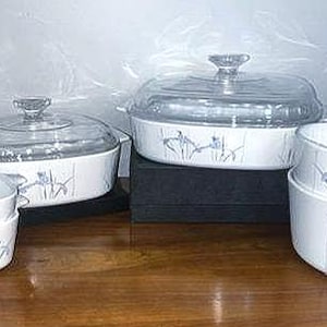 May include: A collection of white ceramic cookware with a floral design. The set includes lidded casserole dishes and handled bowls. The clear glass lids have crystal knobs. The bowls and dishes are stacked on a wooden surface.
