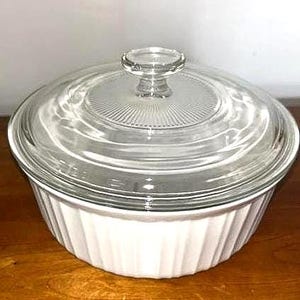 May include: A white, ribbed casserole dish with a clear glass lid. The lid has a central knob and a ridged design. The dish sits on a wooden surface. This is a classic kitchenware item.
