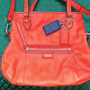 May include: A bright orange leather Coach handbag with a silver zipper and a blue leather tag that says "Coach".