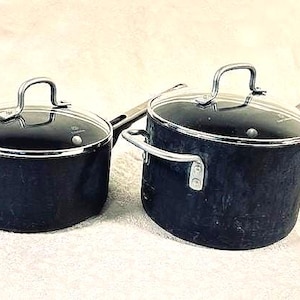 May include: Two black saucepans with glass lids and silver handles. The pans are different sizes, with one having a long handle and the other a shorter handle. The lids have silver rims and handles.