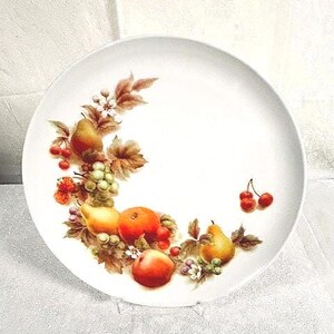 May include: White ceramic plate with a colorful fruit design. The plate features illustrations of pears, cherries, grapes, and oranges, surrounded by leaves and flowers. A decorative piece for serving or display.