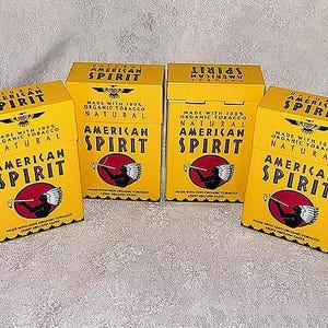 May include: Four yellow boxes of American Spirit cigarettes. Each box features the brand name in bold letters, a Native American figure, and the text "Made with 100% Organic Tobacco Natural". The boxes are arranged on a light-colored surface.