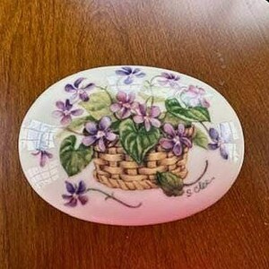 May include: Oval ceramic dish with a hand-painted design of a basket filled with purple violets and green leaves. The basket is a light brown color. The dish has a pink border and is signed "S. Clark".