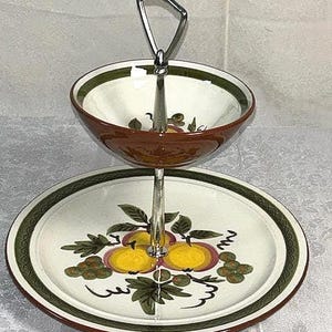 May include: Two-tiered serving dish with a metal handle. The top bowl and bottom plate are white with a brown rim and a green border. The plate features a fruit and leaf design in yellow, green, and brown.