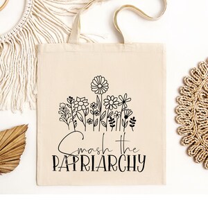 May include: Cream-colored tote bag with a black floral design and the words "Smash the Patriarchy." The bag has a rectangular shape with a handle made of the same material. The design features various flowers.