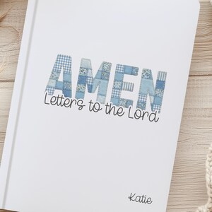 May include: A white notebook with the text "AMEN Letters to the Lord" in blue patchwork lettering. The notebook is personalised with the name "Katie". The image is set on a light wood surface with a cup of coffee and a beige sweater.