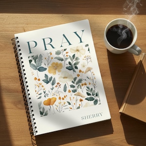 May include: A spiral-bound notebook with a white cover featuring the word PRAY in dark teal letters at the top and the name SHERRY in the bottom right corner. The centre displays a botanical illustration of yellow flowers and teal foliage.