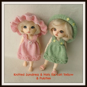 Puede incluir: Dos vestidos de verano de punto y sombreros para muñecas. Los vestidos son rosa y verde, y los sombreros son blancos con flores amarillas. Las muñecas no están incluidas en la imagen.