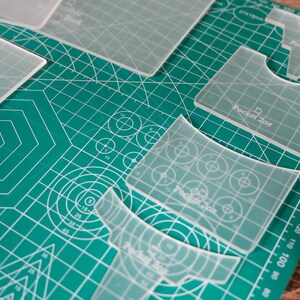 Peut inclure: Gabarits de couture en acrylique transparent, de formes variées, disposés sur un tapis de coupe vert avec des lignes de quadrillage blanches et des mesures. Le texte "Pocket 2oz" est visible.