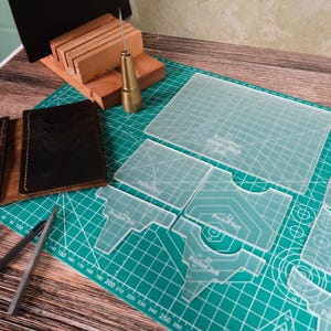 May include: Leathercraft tools and supplies on a green cutting mat. Includes leather wallets, clear acrylic templates, a stitching chisel, and a wooden block. The tools are for crafting leather goods.