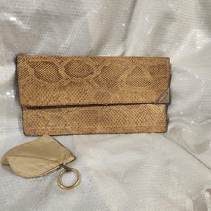 May include: A rectangular tan clutch with a snakeskin texture and a triangular silver-tone metal accent on the right corner. A small, matching tan fabric pouch with a gold-tone metal ring attached by a short strap sits in the lower left corner.