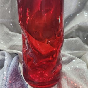 May include: A cylindrical glass tumbler in a vibrant, translucent red hue. The vessel features a distinctively irregular, rippled, and undulating surface texture throughout its body, creating a fluid and organic appearance with light reflections.
