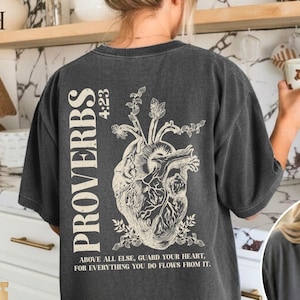 May include: Dark gray t-shirt with a heart and floral design on the back, accompanied by the text "PROVERBS 4:23". The shirt also features the text "ABOVE ALL ELSE, GUARD YOUR HEART, FOR EVERYTHING YOU DO FLOWS FROM IT."