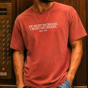 May include: A brick-colored short-sleeve t-shirt featuring the white printed text HE MUST INCREASE, I MUST DECREASE. John 3:30. The shirt has a crew neckline and is worn with a thin silver chain necklace and blue denim jeans.