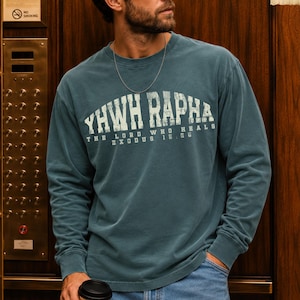 May include: A blue spruce long-sleeve crewneck sweatshirt featuring the white distressed text YHWH RAPHA THE LORD WHO HEALS EXODUS 15:26 printed across the chest. The garment has a relaxed fit with ribbed cuffs and a simple neckline design.