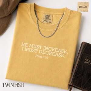 May include: A mustard yellow t-shirt featuring the white text HE MUST INCREASE, I MUST DECREASE. John 3:30 printed on the front. The shirt has a Comfort Colors brand label at the neck, a silver chain necklace resting on the collar, and a brown book nearby.