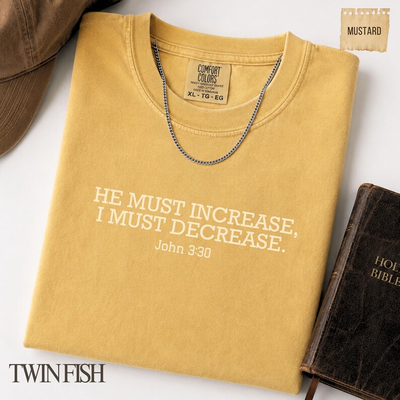 May include: A mustard yellow t-shirt featuring the white text HE MUST INCREASE, I MUST DECREASE. John 3:30 printed on the front. The shirt has a Comfort Colors brand label at the neck, a silver chain necklace resting on the collar, and a brown book nearby.