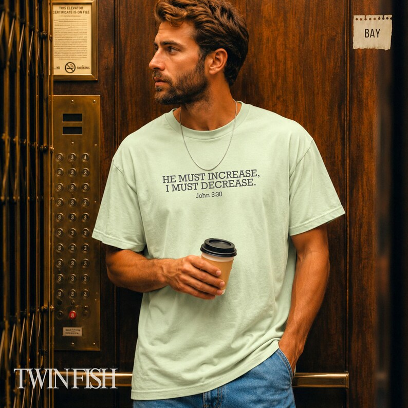 May include: A light green short-sleeve t-shirt featuring the black printed text HE MUST INCREASE, I MUST DECREASE. John 3:30 centered on the chest. The shirt is paired with blue denim jeans and a thin silver chain necklace worn around the neck.