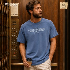 May include: A blue short-sleeved t-shirt featuring the white printed text HE MUST INCREASE, I MUST DECREASE, John 3:30. The shirt has a crew neckline and a relaxed fit. A thin silver chain necklace is worn around the neck.