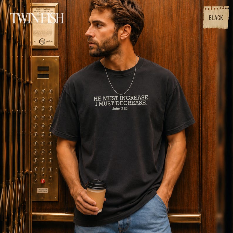 May include: A black short-sleeved t-shirt featuring the white printed text HE MUST INCREASE, I MUST DECREASE. John 3:30 centered on the chest. The shirt has a crew neckline and a standard fit, paired with blue denim jeans and a thin silver chain necklace.
