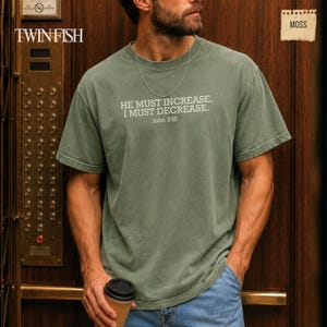 May include: A moss green short-sleeve t-shirt featuring the white printed text "HE MUST INCREASE, I MUST DECREASE. John 3:30" on the chest. The shirt has a crew neckline and a relaxed fit, paired with a thin silver chain necklace.