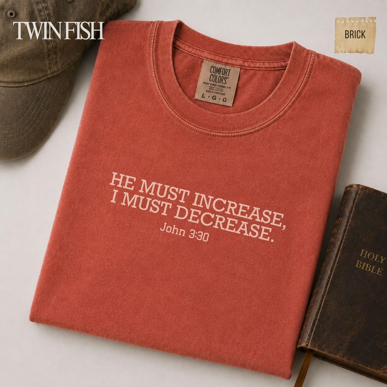 May include: A brick-colored short-sleeve t-shirt featuring the white printed text "HE MUST INCREASE, I MUST DECREASE. John 3:30" on the chest. The shirt has a crew neckline and a Comfort Colors brand label on the inside collar indicating size large.